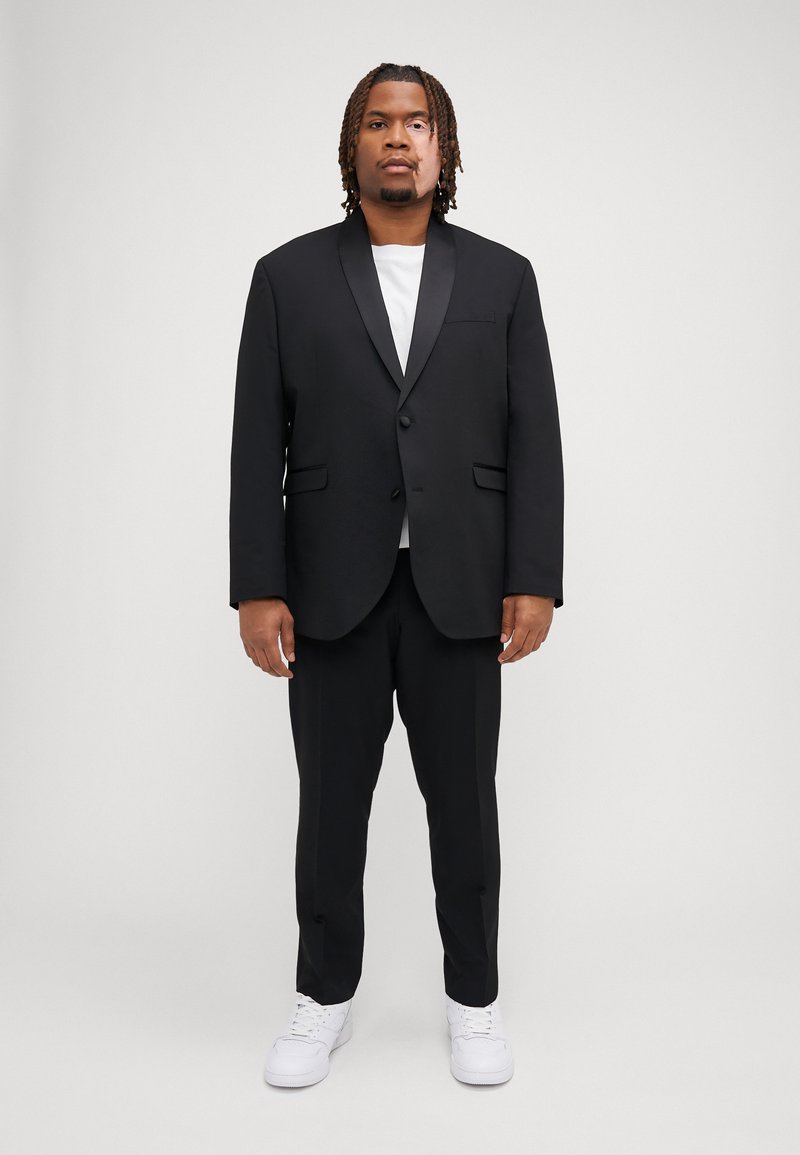 Black suit with a satin peak lapel, paired with a white t-shirt and black trousers, complemented by white sneakers. Formal design with pockets.