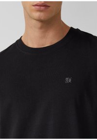 Zbliżenie na mężczyznę w prostym czarnym T-shircie z okrągłym dekoltem, na którym znajduje się małe logo na klatce piersiowej z napisem "s.Ol".