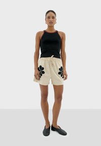 Zwarte mouwloze top en beige shorts met zwarte bloemenborduursels, voorzien van een verstelbare trekkoord taille en een gladde textuur. Zwarte instappers.