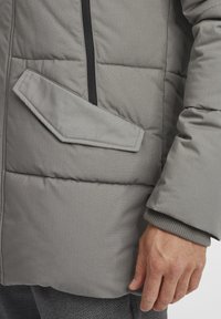 Doudoune grise avec design de poches en angle, tissu texturé et poignets côtelés. Présente une fermeture éclair verticale à l'avant.