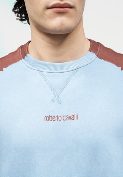 Κοντινό πλάνο ενός ατόμου που φοράει γαλάζιο φούτερ με καφέ λεπτομέρειες στους ώμους και το κείμενο «roberto cavalli» στο στήθος.