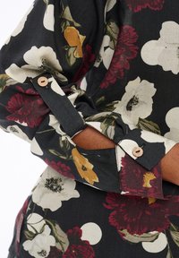 Bras croisés sur le torse, portant une chemise en tissu noir avec un large motif floral blanc, rouge et jaune et des boutons en bois aux poignets.