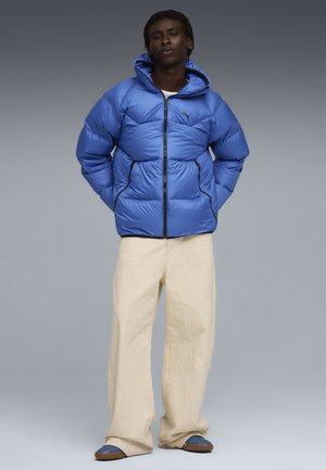Blauwe pufferjack met een capuchon en een ritssluiting, met gestikte vlakken en zwarte accenten. Gecombineerd met crèmekleurige broek en blauwe instapschoenen.