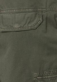 Olijfgroene stof met een gladde textuur, voorzien van een patchzak en contrasterende stiksels voor extra detail.