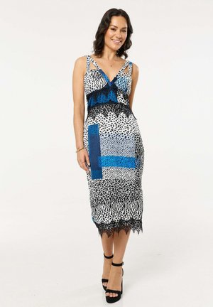 Woman smiling, wearing a sleeveless midi dress with mixed black and blue animal prints, lace details, and black high-heeled sandals.