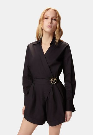 Donna con capelli biondi ondulati che indossa una tuta nera a maniche lunghe con collo incrociato e fibbia decorativa dorata in vita.