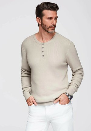 Maglietta henley a maniche lunghe beige a coste, con scollo rotondo e quattro bottoni neri, abbinata a pantaloni bianchi e un orologio argento.