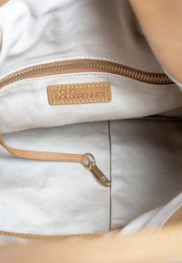 Binnenkant van een tan kleurige tas met een beige voering, voorzien van een gouden rits, een leren label met de tekst "CLIO GOLDBRENNER" en een leren riem bevestiging.