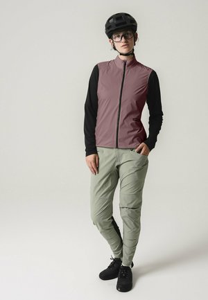 Frau steht und trägt einen schwarzen Helm, ein schwarzes Langarmshirt, eine mauvefarbene ärmellose Weste, salbeigrüne Hosen und schwarze Schuhe vor schlichtem Hintergrund.