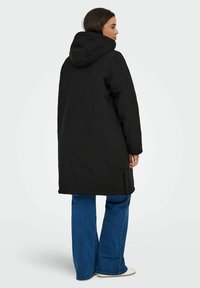 Svart isolerad kappa med huva, långa ärmar och rak skärning. Har sidofickor och en slät, matt yta. Jeans och skor syns inte.