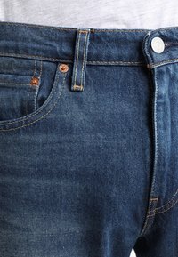 Modré džíny z denimu s měděným knoflíkem a nýty. Mají kapsy a zvýrazněné kontrastní prošití. Materiál je měkký s vybledlou texturou.