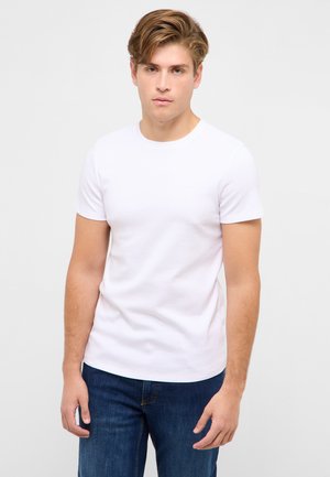 Jeune homme aux cheveux châtain clair debout, portant un t-shirt blanc uni à manches courtes et un jean bleu foncé sur un fond neutre.