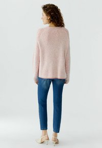 Frau mit lockigem Haar, die einen rosa gestreiften Pullover, blaue enge Jeans und beige Absatzschuhe trägt, steht mit dem Rücken zur Kamera vor einem hellen Hintergrund.
