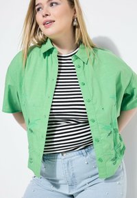Camicia verde a maniche corte con bottoni e ricami a palma, indossata sopra una maglietta a righe bianco e nera; abbinata a pantaloni corti in denim azzurro chiaro decorati con perle.