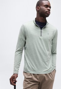 Νεαρός άνδρας με ανοιχτό πράσινο πουλόβερ με φερμουάρ στο λαιμό και μπεζ παντελόνι, κρατώντας μπαστούνι περπατήματος, κοιτάζει προς τα δεξιά σε απλό φόντο.