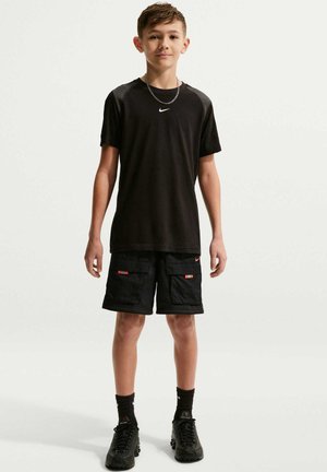 Teenagedreng står iført sort Nike t-shirt, sorte cargo-shorts, sorte sneakers og en sølvkæde om halsen mod en ensfarvet baggrund.