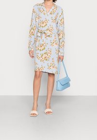 Robe fleurie bleu clair à manches longues, col en V et taille nouée. Associée à des sandales tressées beige et un petit sac à main bleu texturé.