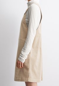 Beige kordfluweel overallklänning med sidofickor, justerbara band och avslappnad passform, bärs över en ljus, långärmad topp.