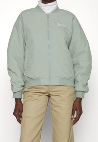 Blouson aviateur vert clair avec poignets et taille élastiques, doté d'une fermeture éclair frontale et d'un petit logo sur la poitrine, porté sur un col roulé blanc.