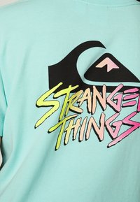 Light blue cotton t-shirt featuring a black logo and multicolored "Stranger Things" text in sharp, cartoonish lettering.