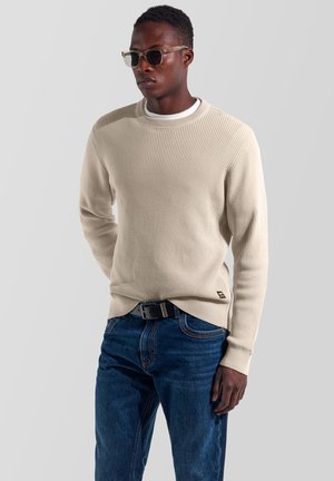 Uomo che indossa un maglione beige texturizzato, jeans blu, cintura nera e occhiali traslucidi, in piedi su sfondo grigio uniforme.