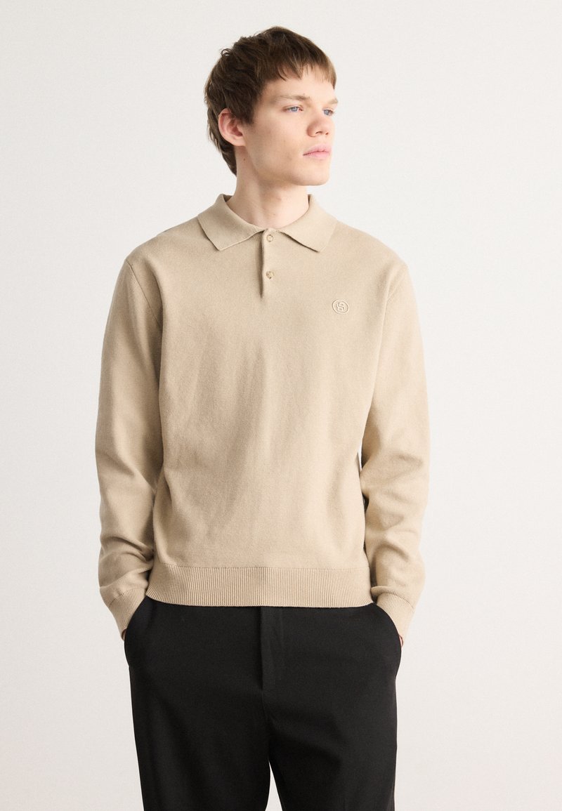 Beige adīts polo džemperis ar garām piedurknēm, ribināta apakša un neliela logo detaļa uz krūtīm. Minimālistisks dizains, mīksta tekstūra.