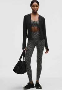 Mujer con chaqueta negra con cremallera, sujetador deportivo gris con estampado de leopardo y leggings, sosteniendo una bolsa de gimnasio negra, de pie sobre un fondo blanco liso.