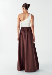 Vrouw met een bun in haar haar, gekleed in een witte top met één schouder en een lange bruine rok, staand met haar rug naar de camera.