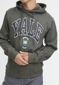 Oliven grøn hoodie med en frontlomme, der har stor hvid tekst "YALE" og et grafisk motiv af et skjold omkranset af laurbærblade.