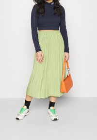 Dunkelblaue, kurz geschnittene Langarmbluse, hellgrüne, plissierte Midirock, orangefarbene Handtasche und Sneakers mit grünen Schnürsenkeln und weißen Akzenten.