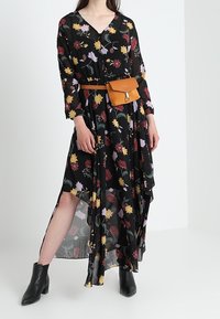 Czarna sukienka w kwiaty z dekoltem w kształcie litery V, długimi rękawami i asymetrycznym dołem; ozdobiona wielokolorowymi wzorami liści i kwiatów; zestawiona z brązową torebką na pasek.