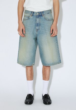 Denim shorts in een lichte blauwe wassing, met een losse pasvorm, steekzakken aan de voorkant en een zoom die tot halverwege de dij komt. Gecombineerd met witte sokken en zwarte schoenen.
