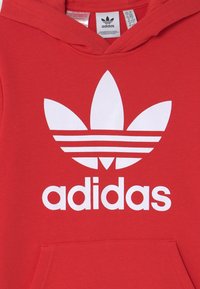 Röd huvtröja i bomull med en stor vit Adidas-logotyp och tre horisontella ränder ovanför ett lövliknande mönster.