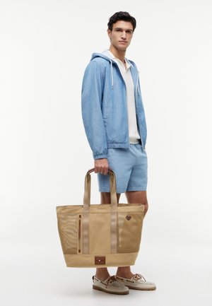 Mand iført lyseblå jakke og shorts, der holder en stor beige shopper, stående mod en hvid baggrund.