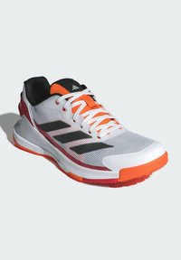 Witte sportieve schoen met oranje accenten, zwarte binnenzool en bovenwerk van mesh. Beschikt over drie zwarte strepen en een gestructureerde rubberen zool.