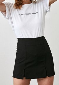 Mujer vestida con una camiseta blanca que dice "QUIERO UN BILLETE A CUALQUIER LUGAR" y una falda corta negra de tiro alto con aberturas frontales, de pie contra una pared blanca.