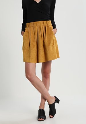 Jupe patineuse en suédine jaune moutarde avec une taille élastique, des poches latérales et une silhouette évasée, assortie à des mules noires à bout ouvert.
