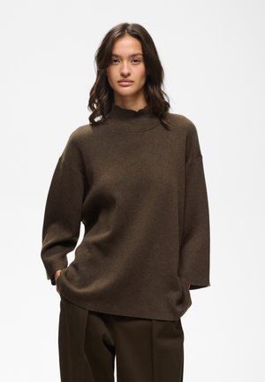 Femme aux cheveux foncés ondulés portant un pull marron oversize et un pantalon marron assorti, debout les mains dans les poches devant un fond uni.