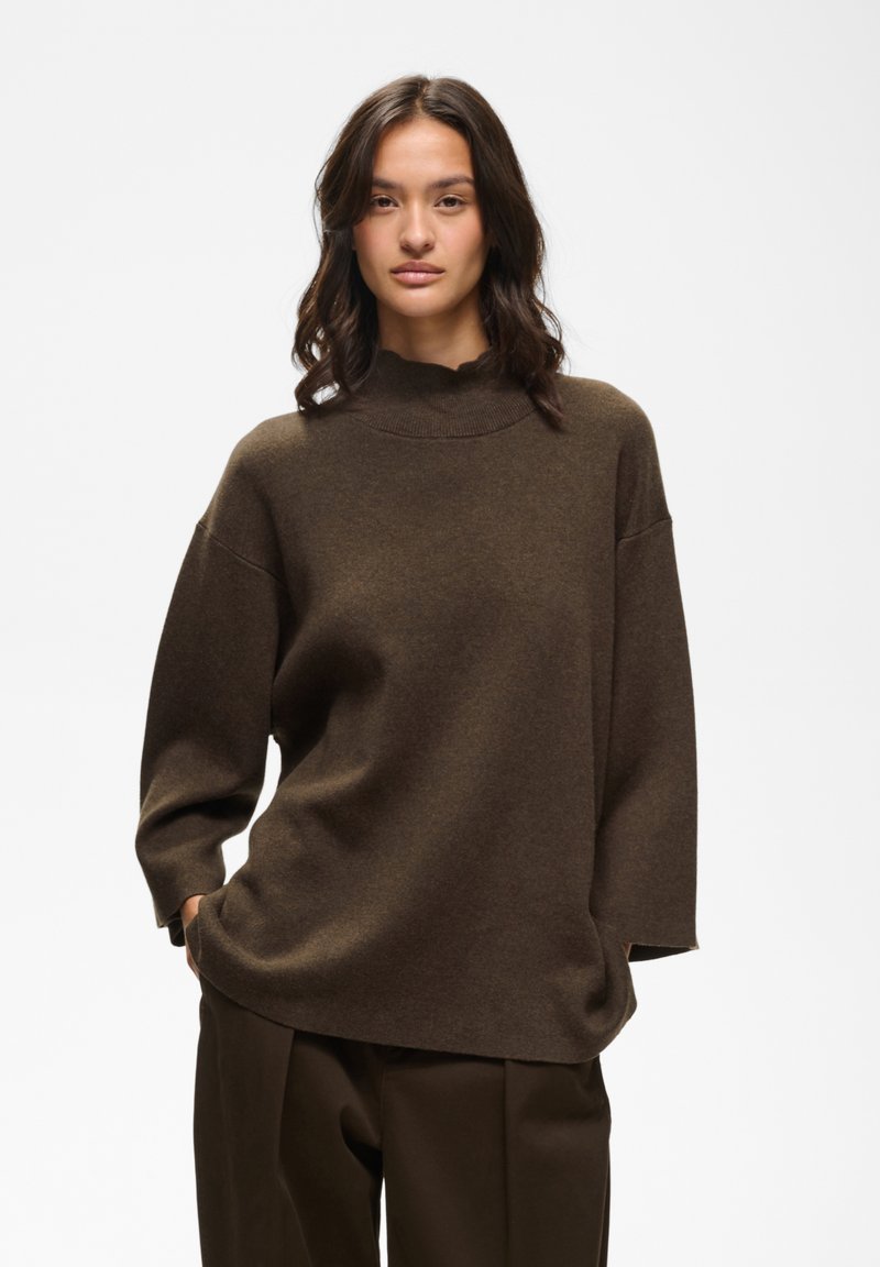 Femme aux cheveux foncés ondulés portant un pull marron oversize et un pantalon marron assorti, debout les mains dans les poches devant un fond uni.