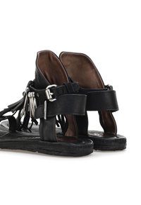 Un par de sandalias de cuero negro con correas para el tobillo, hebilla plateada y detalles de flecos, mostradas desde atrás sobre un fondo blanco.