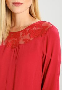 Blusa rossa con yoke in pizzo floreale, scollatura rotonda e maniche lunghe. Tessuto morbido con una texture liscia.