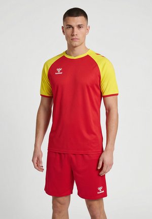 Mann steht, trägt ein rot-gelbes Sporttrikot und rote Shorts mit hummel-Logos auf dem Shirt und den Shorts.