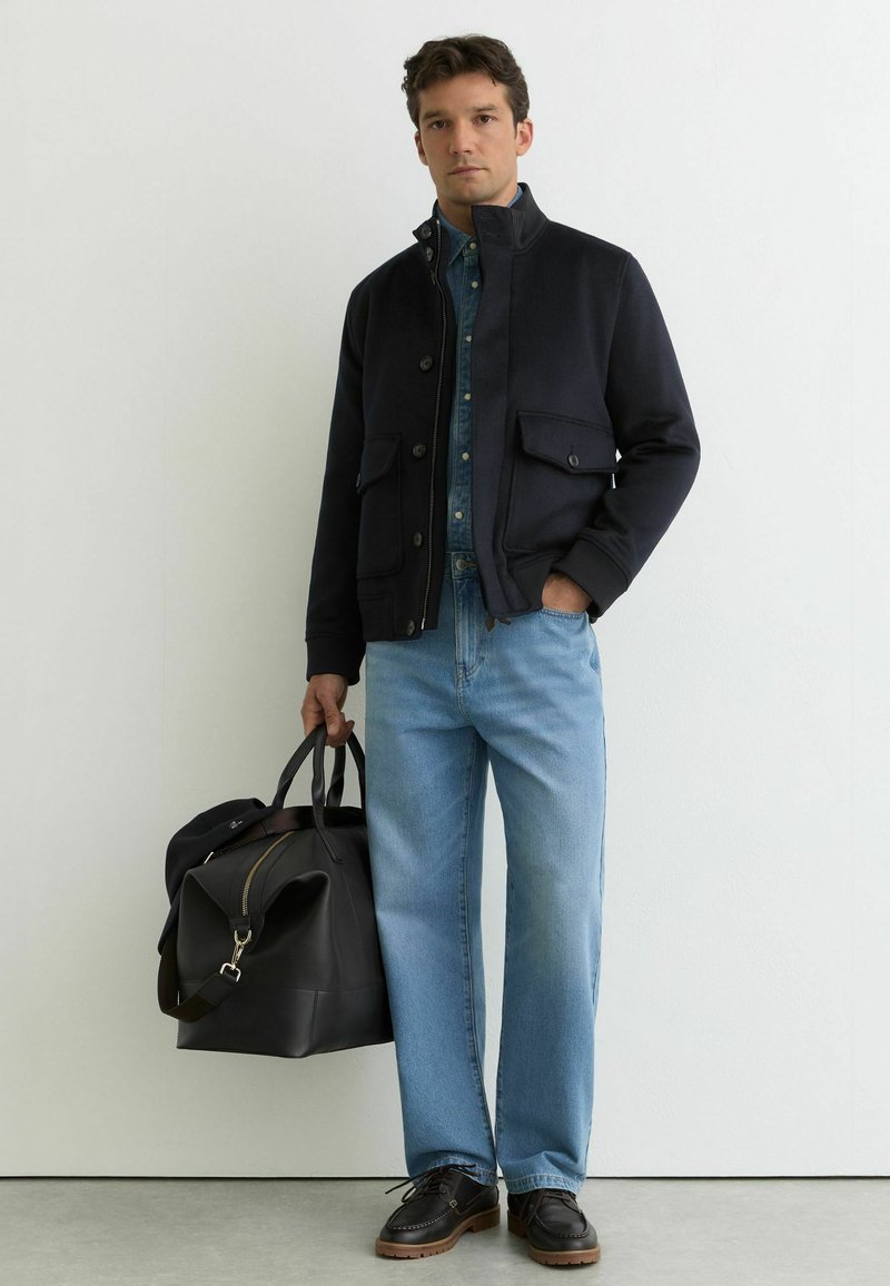 Mann trägt eine schwarze Jacke, blaues Hemd mit Knöpfen, hellblaue Jeans und schwarze Schuhe, hält eine große schwarze Lederreisetasche.