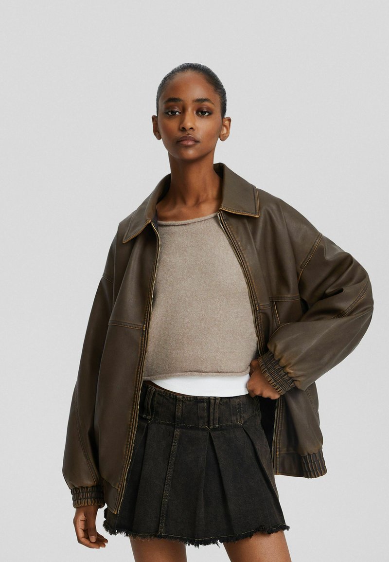 Chaqueta de cuero marrón con un gran cuello y puños acanalados, combinada con un suéter corto beige y una falda negra con pliegues.