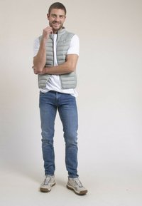 Veste matelassée gris clair sur un t-shirt blanc, associée à un jean bleu et des baskets beiges. La veste comporte une fermeture éclair et un logo.