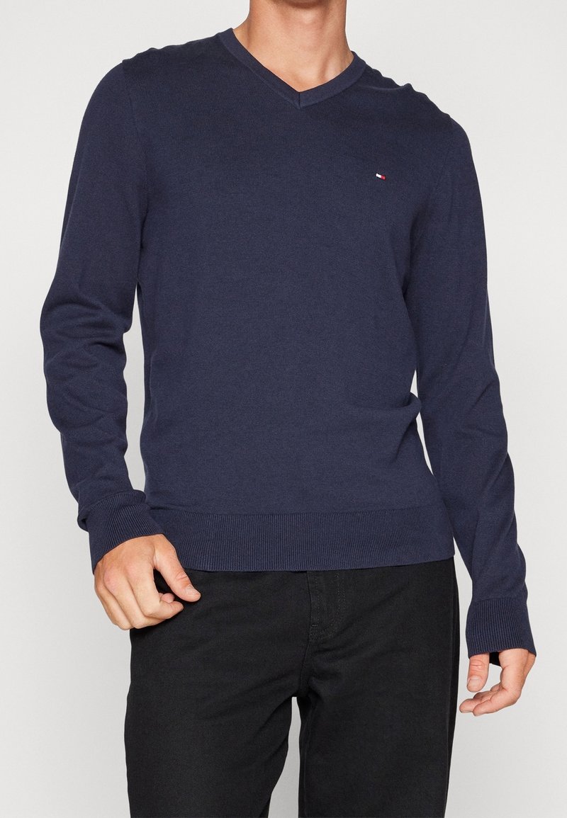 Homme portant un pull bleu marine à col en V avec manches longues et petit logo sur la poitrine, assorti à un pantalon noir.