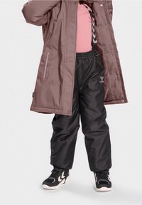 Veste matelassée marron, pantalon imperméable noir et baskets noires avec des accents blancs. La tenue présente un design simple et une taille élastiquée.