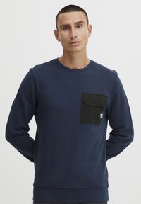 Mørkeblå sweatshirt med lange ermer, rund hals og en svart lomme på venstre bryst. Har ribbestrikket falde og mansjetter.