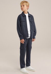 Tuta blu navy realizzato in tessuto testurizzato, composto da una giacca con zip e due tasche frontali, un paio di pantaloni cargo e scarpe da ginnastica bianche.