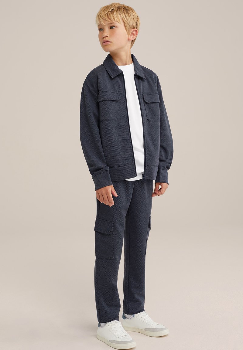 Tuta blu navy realizzato in tessuto testurizzato, composto da una giacca con zip e due tasche frontali, un paio di pantaloni cargo e scarpe da ginnastica bianche.